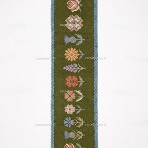 May include: A long, narrow, olive green rug with a blue border and white fringe. The rug features a repeating pattern of stylized flowers in various colors, including pink, orange, and blue. The floral design is centered on the rug's length.