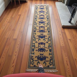 Op de afbeelding: Een rechthoekige loper met een beige achtergrond en blauwe en marineblauwe geometrische patronen. De loper heeft aan beide uiteinden franjes en ligt op een houten vloer. De loper is een decoratief element voor een gang of entree.