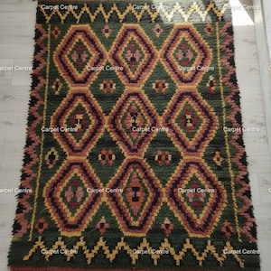 May include: A rectangular, hand-woven rug with a geometric diamond pattern in shades of green, burgundy, and gold. The rug has a fringed edge and a zig-zag border. The text "Carpet Centre" is repeated across the rug.