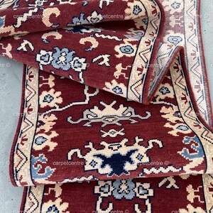 Puede incluir: Una alfombra tipo pasillo con fondo rojo intenso y un patrón repetitivo de motivos florales y geométricos estilizados en crema, azul claro y azul marino. La alfombra tiene un borde ancho con diseño geométrico crema y un borde rojo oscuro.