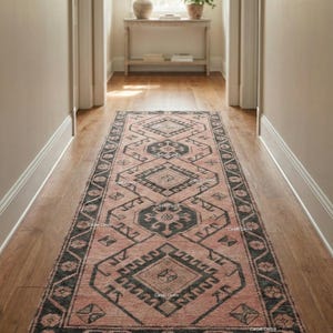 May include: A pink and charcoal grey runner rug with a geometric pattern, laid on a wooden floor in a hallway. The rug's design includes diamond and floral shapes. The rug is a long, rectangular shape.