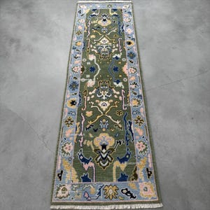 May include: A long, rectangular olive green rug with an intricate floral pattern. The rug features a light blue border with pink and yellow floral accents. The central design includes navy blue and yellow details. The rug is placed on a gray surface.