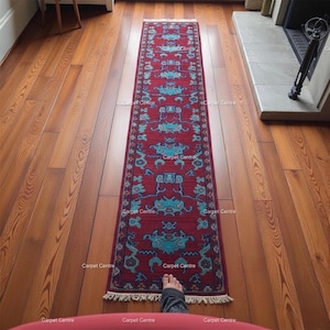 Op de afbeelding: Een lange, rechthoekige rode loper met een herhalend turquoise patroon en franjes. De loper ligt op een houten vloer. De woorden "Carpet Centre" staan op de loper. De loper is ongeveer 200 cm lang.