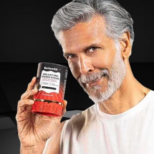 May include: A cylindrical container of "BetterAlt Shilajit Pro Energy Sticks" held by a person. The container is primarily red and black with white text. The person is wearing a white shirt and has gray hair and a beard.