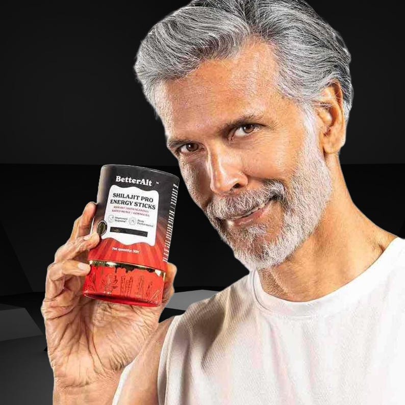 May include: A cylindrical container of "BetterAlt Shilajit Pro Energy Sticks" held by a person. The container is primarily red and black with white text. The person is wearing a white shirt and has gray hair and a beard.