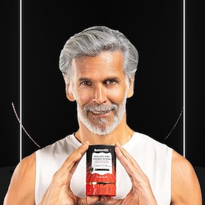 May include: A man with gray hair and a beard holds a cylindrical product labeled "BetterAlt Shilajit Pro Energy Sticks." The product packaging is black, red, and gold. The background is black with white lines.