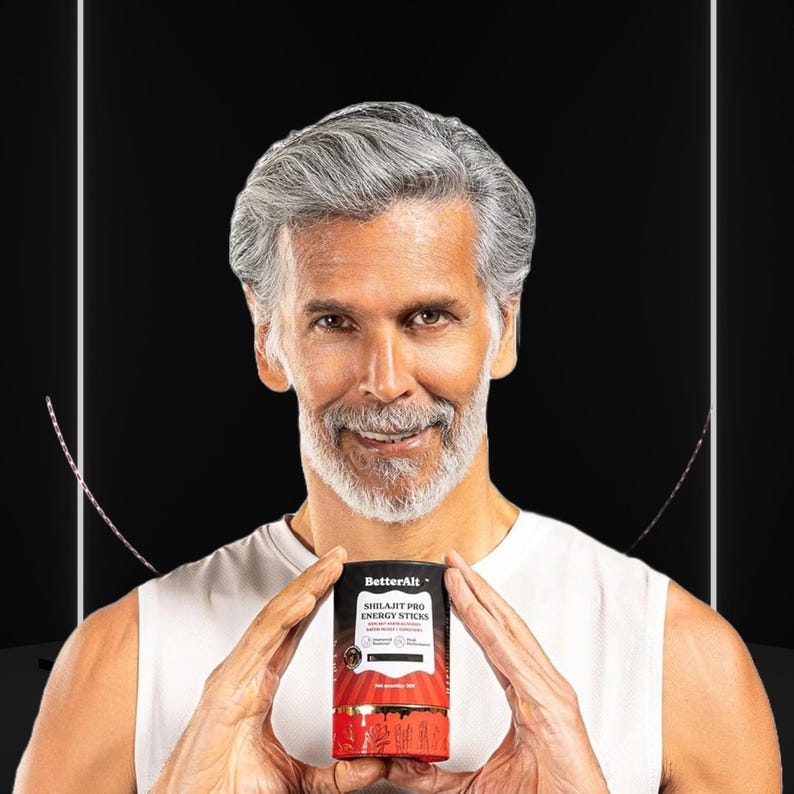 May include: A man with gray hair and a beard holds a cylindrical product labeled "BetterAlt Shilajit Pro Energy Sticks." The product packaging is black, red, and gold. The background is black with white lines.