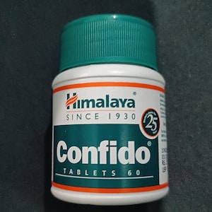May include: A white bottle of Himalaya Confido tablets with a green cap. The label features the Himalaya logo, the word "Confido" in a bold font, and the text "TABLETS 60". The bottle has an orange stripe.