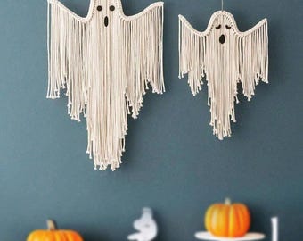 May include: Two macrame ghost wall hangings with open mouths and eye holes. The ghosts are cream-colored and have long fringe. Below are several orange pumpkins of varying sizes, perfect for Halloween decor.
