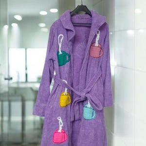 Könnte beinhalten: Ein lila Frottee-Bademantel mit Kapuze und Bindegürtel. Der Bademantel ist mit gestickten Kaffeetassen-Motiven in Grün, Gelb, Rosa und Blau verziert. Er hängt an einem schwarzen Kleiderbügel.
