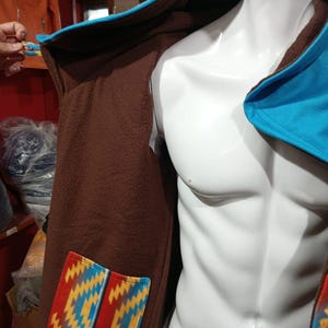 May include: Brown hooded coat with a blue trim and a colorful patterned pocket. The coat is displayed on a white mannequin. The pocket features a geometric design in red, yellow, and blue.