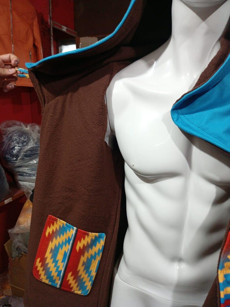 May include: Brown hooded coat with a blue trim and a colorful patterned pocket. The coat is displayed on a white mannequin. The pocket features a geometric design in red, yellow, and blue.