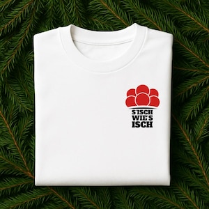 Könnte beinhalten: Weißes T-Shirt mit Rundhalsausschnitt und kleinem Aufdruck auf der Brust. Die Grafik zeigt eine Ansammlung roter Formen über dem Text "S'ISCH WIE'S ISCH" in Schwarz. Das T-Shirt wird auf einem Hintergrund aus grünen Tannennadeln präsentiert.