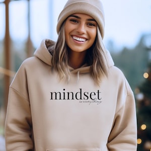Könnte beinhalten: Beigefarbener Kapuzenpullover mit dem Text "mindset is everything" in Schwarz. Der Kapuzenpullover hat eine Vordertasche und eine Kapuze mit Kordelzug. Die Person auf dem Bild trägt eine beige Mütze.