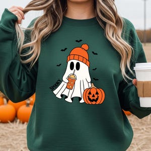 May include: Dark green sweatshirt featuring a cartoon ghost wearing an orange beanie and holding a pumpkin spice drink. The ghost is next to an orange jack-o'-lantern. The word "Spooky" is printed on the sweatshirt. Black bats are in the background.