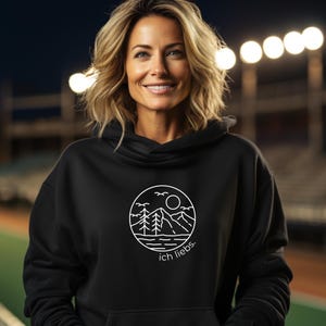 May include: Black hooded sweatshirt with a white circular graphic featuring a mountain range, trees, sun, and birds. The text "ich liebs." is below the graphic. The sweatshirt has a drawstring hood.