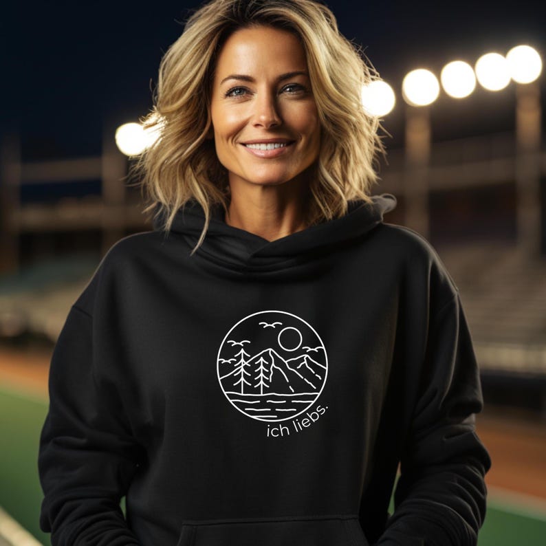 May include: Black hooded sweatshirt with a white circular graphic featuring a mountain range, trees, sun, and birds. The text "ich liebs." is below the graphic. The sweatshirt has a drawstring hood.