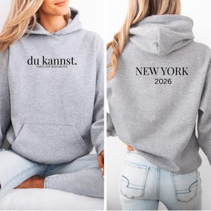 Könnte beinhalten: Ein grauer Kapuzenpullover mit einer Vordertasche und einer Kapuze. Auf der Vorderseite des Kapuzenpullovers steht in schwarzer Schrift "du kannst. ENDE DER GESCHICHTE". Auf der Rückseite steht in schwarzer Schrift "NEW YORK 2026".