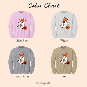 May include: A color chart displays four crewneck sweatshirts in light pink, white, sport grey, and sand. Each sweatshirt features a ghost graphic wearing a beanie and holding a drink, with a pumpkin. The text "Color Chart" is at the top.