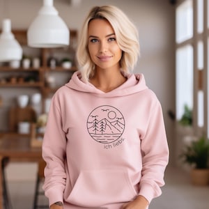 May include: A light pink hoodie with a black circular graphic featuring a mountain landscape, trees, sun, and birds. The text "ich liebs" is below the graphic. The hoodie has a front pocket and a hood.