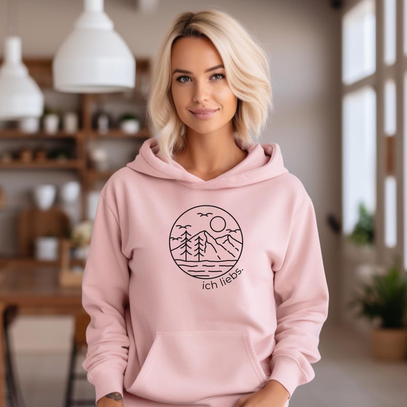 May include: A light pink hoodie with a black circular graphic featuring a mountain landscape, trees, sun, and birds. The text "ich liebs" is below the graphic. The hoodie has a front pocket and a hood.