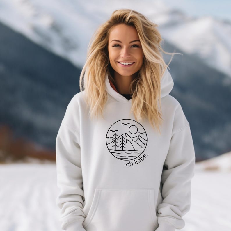 May include: White hoodie with a black circular graphic featuring a mountain scene, trees, sun, and the text "ich liebs." The hoodie has a hood and a front pocket. The person in the image is smiling.