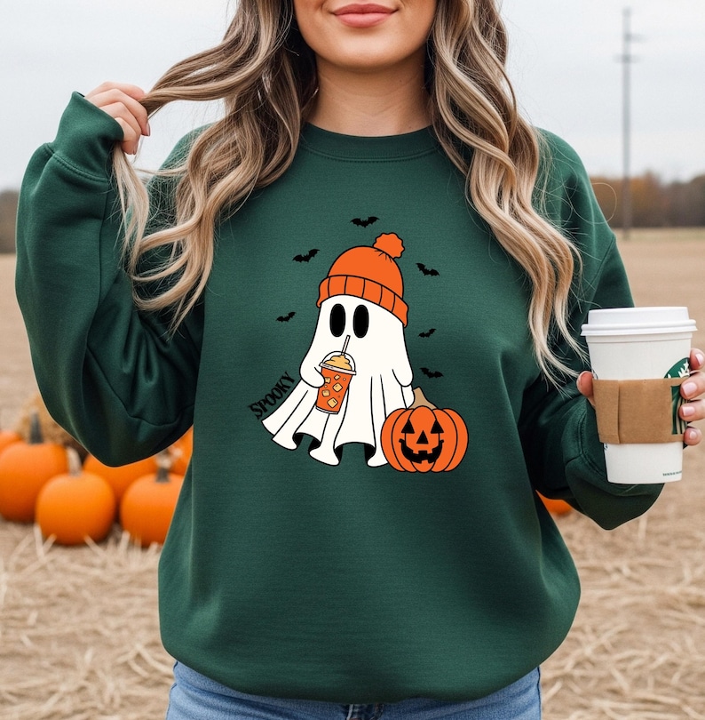 May include: Dark green sweatshirt featuring a cartoon ghost wearing an orange beanie and holding a pumpkin spice drink. The ghost is next to an orange jack-o'-lantern. The word "Spooky" is printed on the sweatshirt. Black bats are in the background.