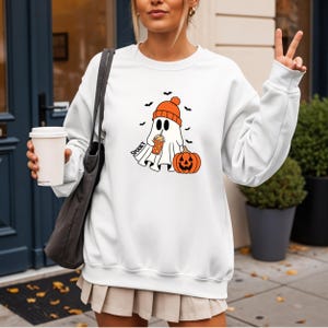 May include: White sweatshirt with a Halloween-themed graphic. The design features a ghost wearing an orange beanie, holding a pumpkin spice drink, and a jack-o'-lantern. The word "Spooky" is printed on the design. Black bats are also included in the design.