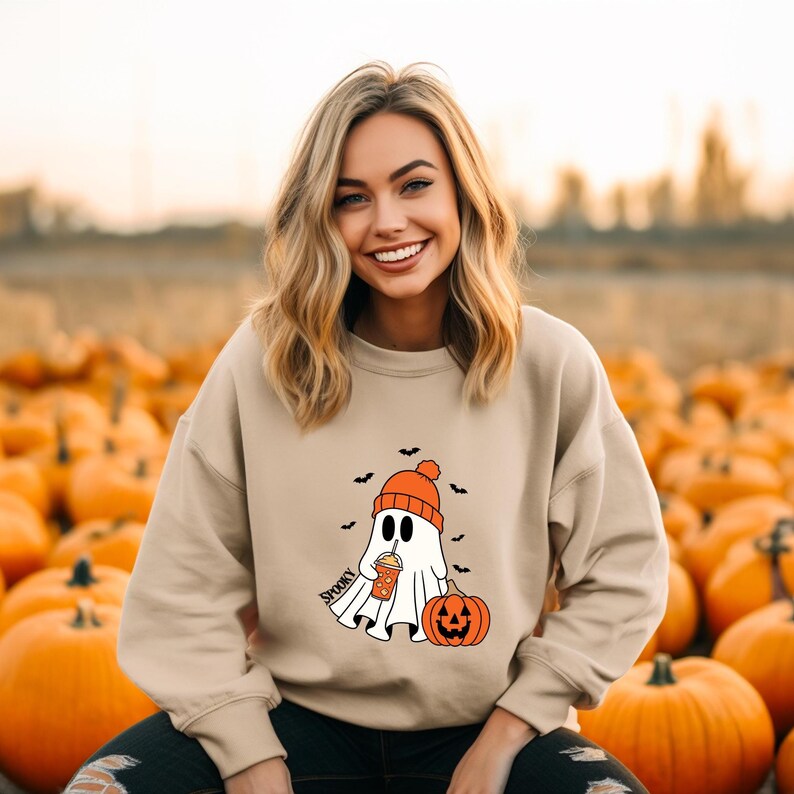 May include: Beige sweatshirt featuring a cartoon ghost wearing an orange beanie and holding a coffee cup, with a jack-o'-lantern and bats. The word "Spooky" is printed on the sweatshirt. The background is a pumpkin patch.