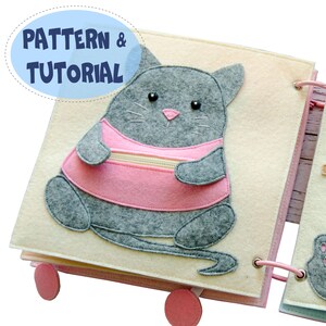 Könnte beinhalten: Ein handgefertigtes Filz-Quiet-Book mit einem grauen Katzendesign, das ein rosa Hemd trägt. Das Buch ist geöffnet und zeigt Seiten mit einer Katzenapplikation. Der Text "PATTERN & TUTORIAL" befindet sich in einem blauen Oval. Das Buch ist mit rosa Kordel gebunden.