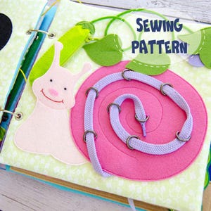 May include: A handmade felt quiet book page featuring a pink snail with a purple shoelace trail. The page includes green felt leaves and a green felt vegetable. The text "SEWING PATTERN" is displayed in blue.