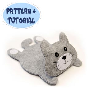 May include: A gray felt cat-shaped craft with white paw pads and a white face. The cat has black eyes and whiskers. The text "PATTERN & TUTORIAL" is in a blue oval above the cat.