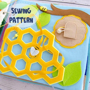 Puede incluir: Un patrón de costura de fieltro azul con un panal amarillo, abejas y las palabras "SEWING PATTERN". El libro presenta una abeja de fieltro, una colmena de fieltro y hojas de fieltro verdes. Una cinta amarilla está enhebrada a través del panal.