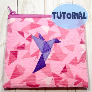 May include: A pink zippered pouch with a geometric patchwork design. A purple origami bird is appliqued on the front. The word "TUTORIAL" is in a blue oval in the upper right corner. The zipper and trim are a darker pink.
