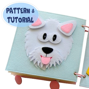 May include: A handmade felt quiet book with a light blue cover featuring a white dog face with pink accents. The book has pink binding and wheels, and the text "PATTERN & TUTORIAL" is in a blue oval.
