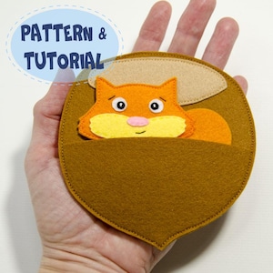 May include: A felt acorn-shaped pouch with an orange squirrel peeking out. The squirrel has a yellow belly and pink nose. The pouch is brown with a tan top. Text in a blue oval reads "PATTERN & TUTORIAL".