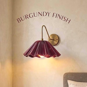 May include: A wall-mounted lamp featuring a burgundy, fluted, bell-shaped shade and a brass-toned arm and circular wall plate. The text BURGUNDY FINISH is printed in an arc above the lamp, which emits a warm light from a central bulb.
