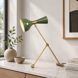 May include: A table lamp with a dual-cone olive green shade and a gold-toned metal frame. The lamp features an adjustable arm structure with a tripod-style base, a spherical end cap, and a cylindrical base support resting on a flat surface.