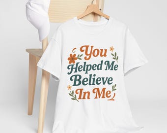 Könnte beinhalten: Ein weißes T-Shirt mit dem Text "You Helped Me Believe In Me" in Orange und Blaugrün, verziert mit floralen Akzenten. Eine weiße Baseballkappe liegt auf einem Holzstuhl im Hintergrund.