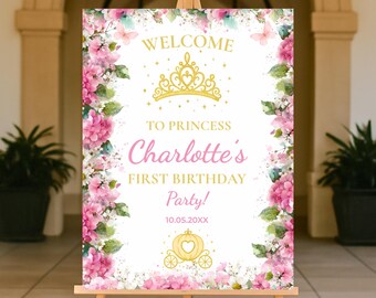 May include: A wooden easel displays a welcome sign for a princess's first birthday party. The sign features a gold crown, pink flowers, and the text "Welcome to Princess Charlotte's First Birthday Party!" with a date and a carriage illustration.