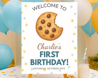 May include: A wooden easel displays a sign with the text "Welcome to Charlie's First Birthday!" and a graphic of a bitten chocolate chip cookie. The sign is surrounded by blue and yellow balloons, with cookies and a milk bottle in the foreground.