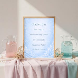 May include: A wooden-framed sign with the text "Glacier Bar" and a list of drink options. Two glass pitchers, one with pink liquid and the other with blue liquid, flank the sign. The table is draped with pink and blue fabric.