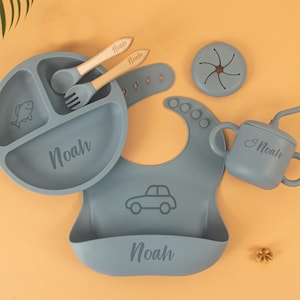 Könnte beinhalten: Ein Baby-Ess-Set in gedämpftem Blau, einschließlich einer geteilten Platte mit Fischmuster, einer Lätzchen mit Autografik, einem Becher mit Strohhalm, einem Snackbehälter sowie einem Löffel und einer Gabel. Der Name "Noah" ist aufgedruckt.