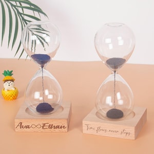 Puede incluir: Dos relojes de arena de cristal transparente con arena azul, sobre bases de madera. Una base está grabada con "Ava ∞ Ethan", la otra con "Time flows, never stops". Una pequeña figura de piña a la izquierda.
