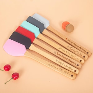 Könnte beinhalten: Eine Auswahl an farbigen Silikon-Teigschabern mit Holzgriffen. Jede Teigschaber hat einen unterschiedlich farbigen Kopf, darunter rosa, schwarz, rot, türkis, grau und hellgrau. Die Holzgriffe sind mit Sätzen wie "Cooking Through the Years" graviert.