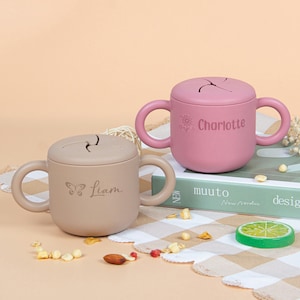 Könnte beinhalten: Zwei Silikon-Snackbecher mit Deckel und Griffen. Einer ist beige mit "Liam" und einem Schmetterling, der andere ist rosa mit "Charlotte" und einer Blume. Beide haben einen Schlitz im Deckel.