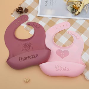 May include: Two silicone baby bibs, one mauve and one pink, with curved necklines and food catchers. The mauve bib features a flower design and the name "Charlotte." The pink bib has a heart and the name "Olivia." Both have adjustable closures.