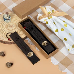 Könnte beinhalten: Zwei dunkle Holzlesezeichen mit goldenen Fächermotiven und dem Text "Life is short and reading is long" und "A book is a dream that you hold in your hand." Ein Lesezeichen befindet sich in einer Schachtel. Eine kleine Keramikfigur und Eicheln sind ebenfalls vorhanden.