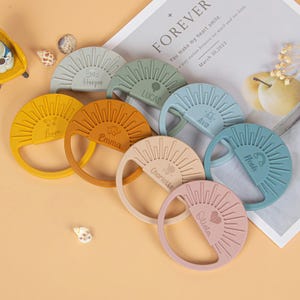 Op de afbeelding: Een verzameling kleurrijke, ronde bijtringen voor baby's in verschillende pasteltinten, elk met een zonnesterontwerp en een naam. De bijtringen zijn gemaakt van zacht, flexibel materiaal en zijn ontworpen voor baby's. Namen zijn Liam, Emma, Charlotte en Olivia.
