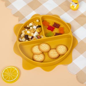 Könnte beinhalten: Ein gelber Teller aus Silikon mit drei Fächern in Form einer Sonne. Der Teller enthält Cracker, Trockenfrüchte und Gummibärchen. Ein gelber Untersetzer in Zitronenform und ein Ananas-Spielzeug sind ebenfalls zu sehen.